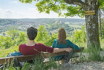 Altmühltal-Panoramaweg (Eichstätt) Zwei Personen sitzen auf einer Bank unter Baum mit Schild „Hohes Kreuz“ und blicken auf ein Tal mit Stadt und Hügeln.
